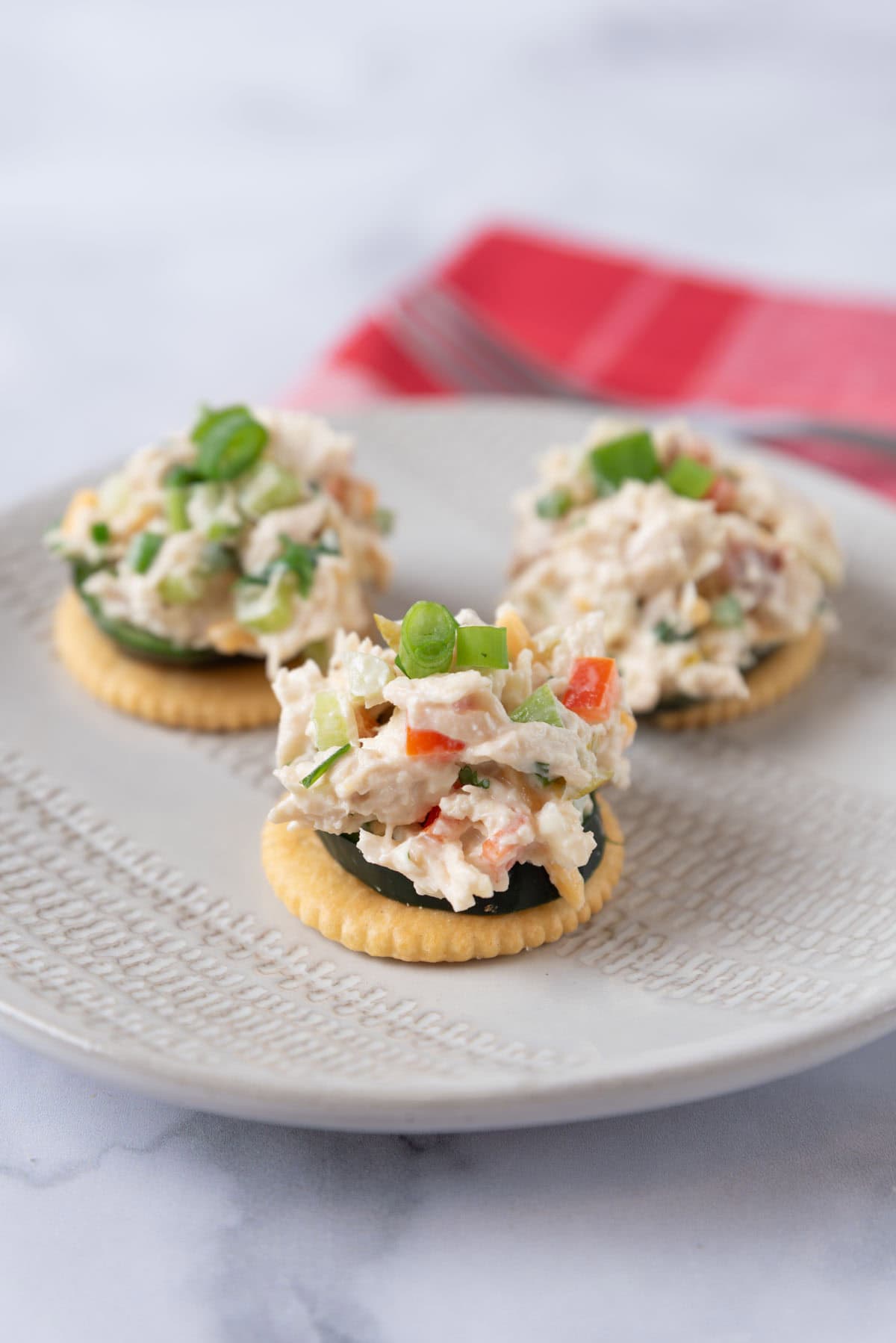 A cream colored plate with three appetizers of a butter cracker stacked with a slice of cucumber and a scoop of jalapeno chicken salad. The tops are garnished with extra green onion. A red linen napkin is out of focus behind the plate.