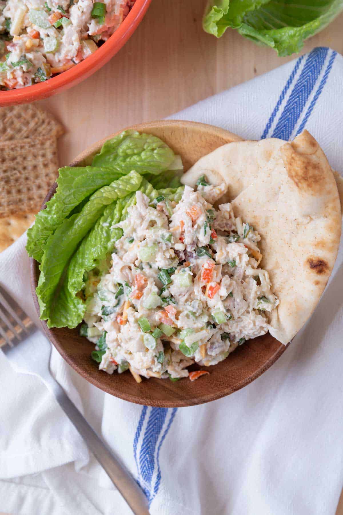 A big scoop of jalapeno chicken salad in a wooden bowl with lettuce leaves and pita wedges nestled in around the edges of the bowl. A wooden board under the bowl with a blue and white linen napkin, wheat crackers more lettuce leaves and the corner of the larger serving bowl in the corner. A fork rests to the left of the bowl.