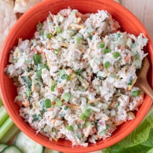 A closeup of a bright orange bowl loaded with Jalapeno Chicken Salad with a wooden spoon in it. Flecks of cheese, peppers, cilantro and green onion visible in the salad. The bowl rests on a wooden board along with pita bread, lettuce leaves, and sliced veggies.
