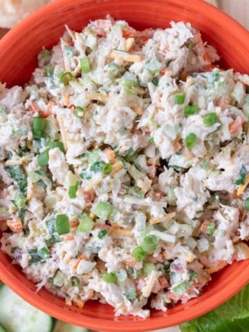 A closeup of a bright orange bowl loaded with Jalapeno Chicken Salad with a wooden spoon in it. Flecks of cheese, peppers, cilantro and green onion visible in the salad. The bowl rests on a wooden board along with pita bread, lettuce leaves, and sliced veggies.