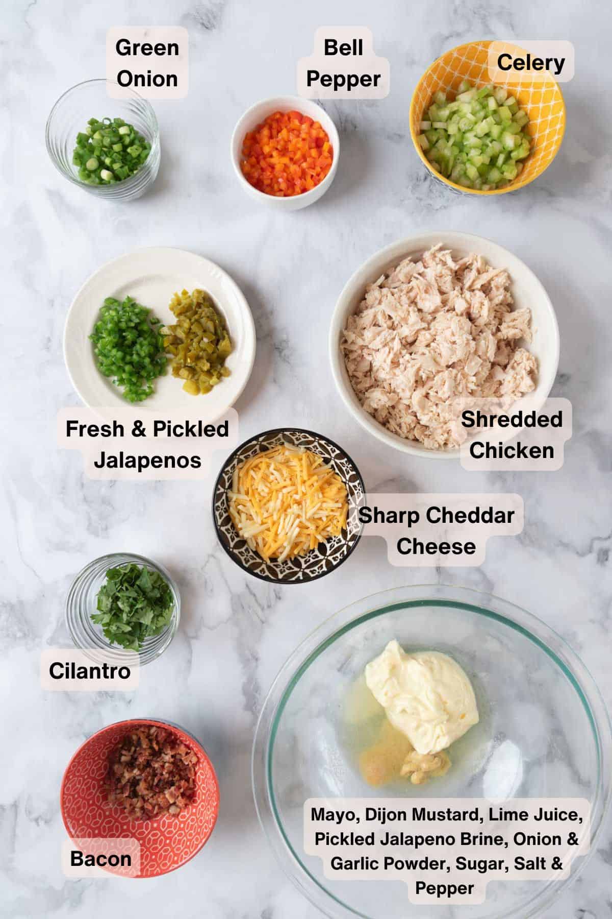 Ingredients for jalapeno chicken salad in various small bowls and dishes scattered around a white and grey marble countertop.