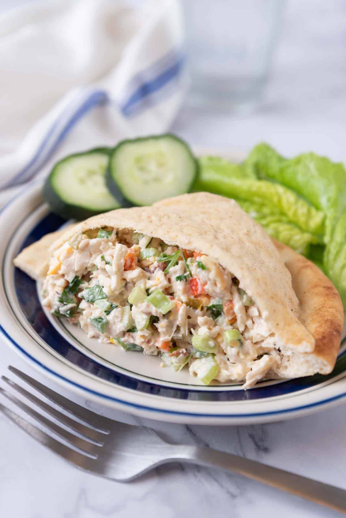 A pita pocket stuffed with Jalapeno Chicken Salad on a white plate with a blue rim on a white marble countertop. A fork rests just in front of the plate.