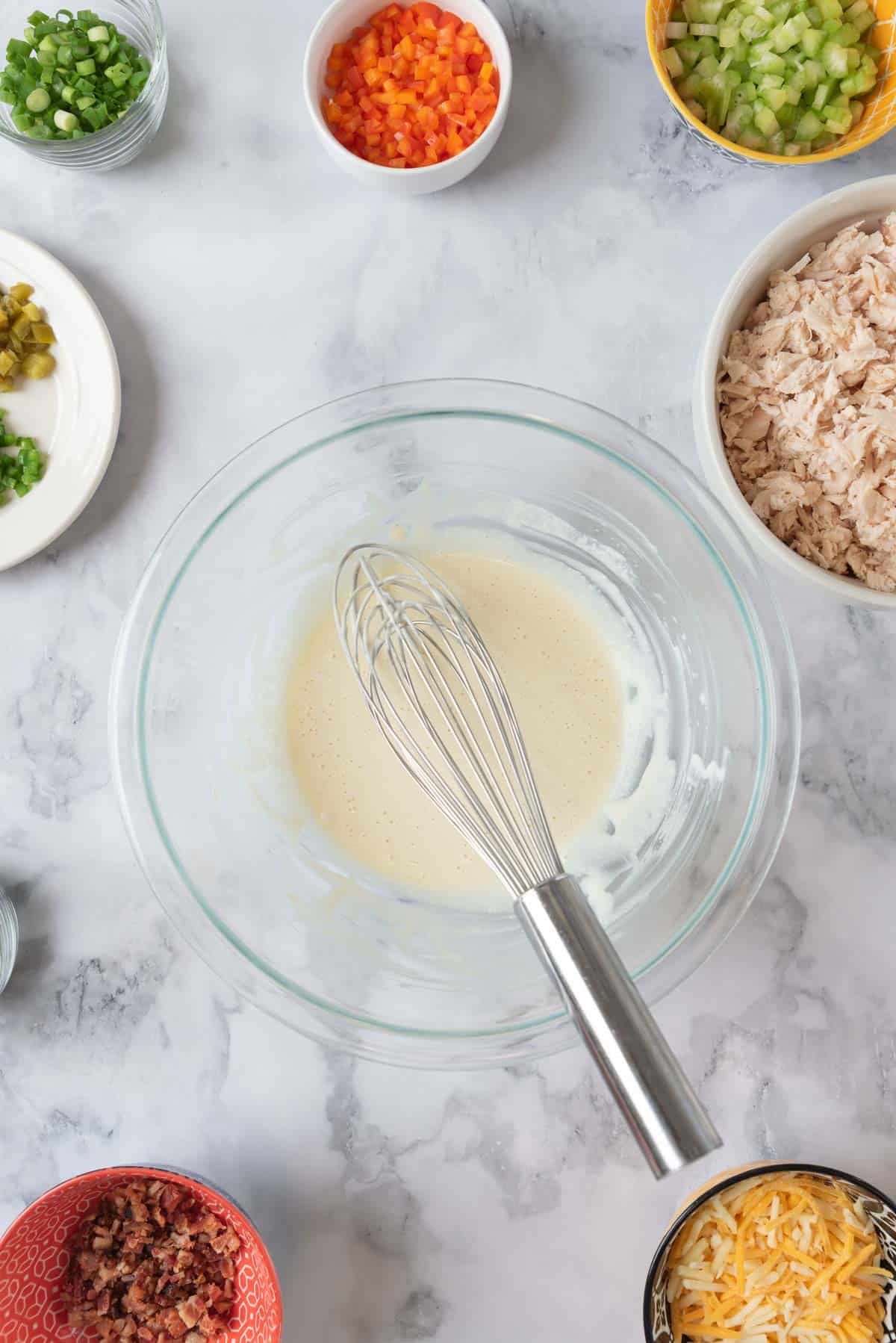 A mixing bowl with salad dressing whisked together. Around the edges are smaller bowls with prepped ingredients.