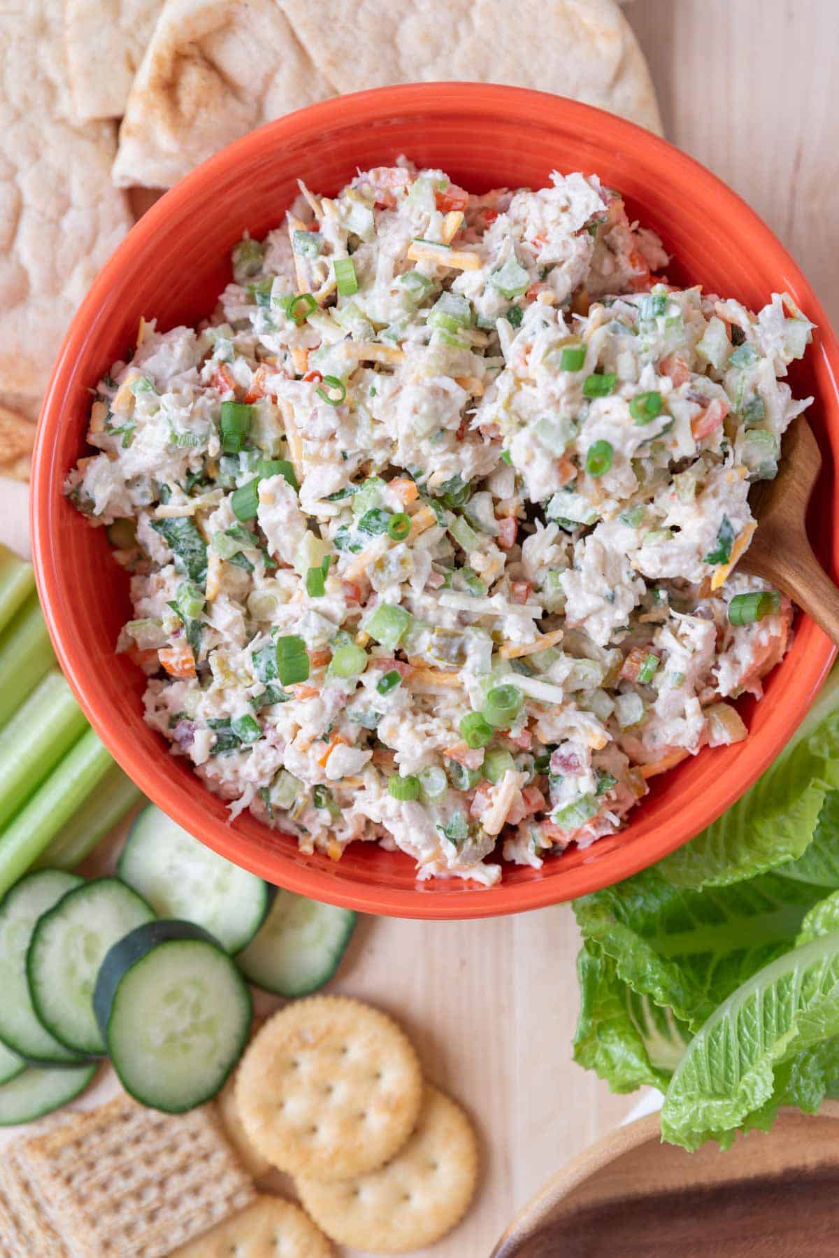 A bright orange bowl filled with Jalapeno Chicken Salad with a wooden spoon in it. Flecks of cheese, peppers, cilantro and green onion visible in the salad. The bowl rests on a wooden board along with pita bread, lettuce leaves, crackers and sliced veggies.