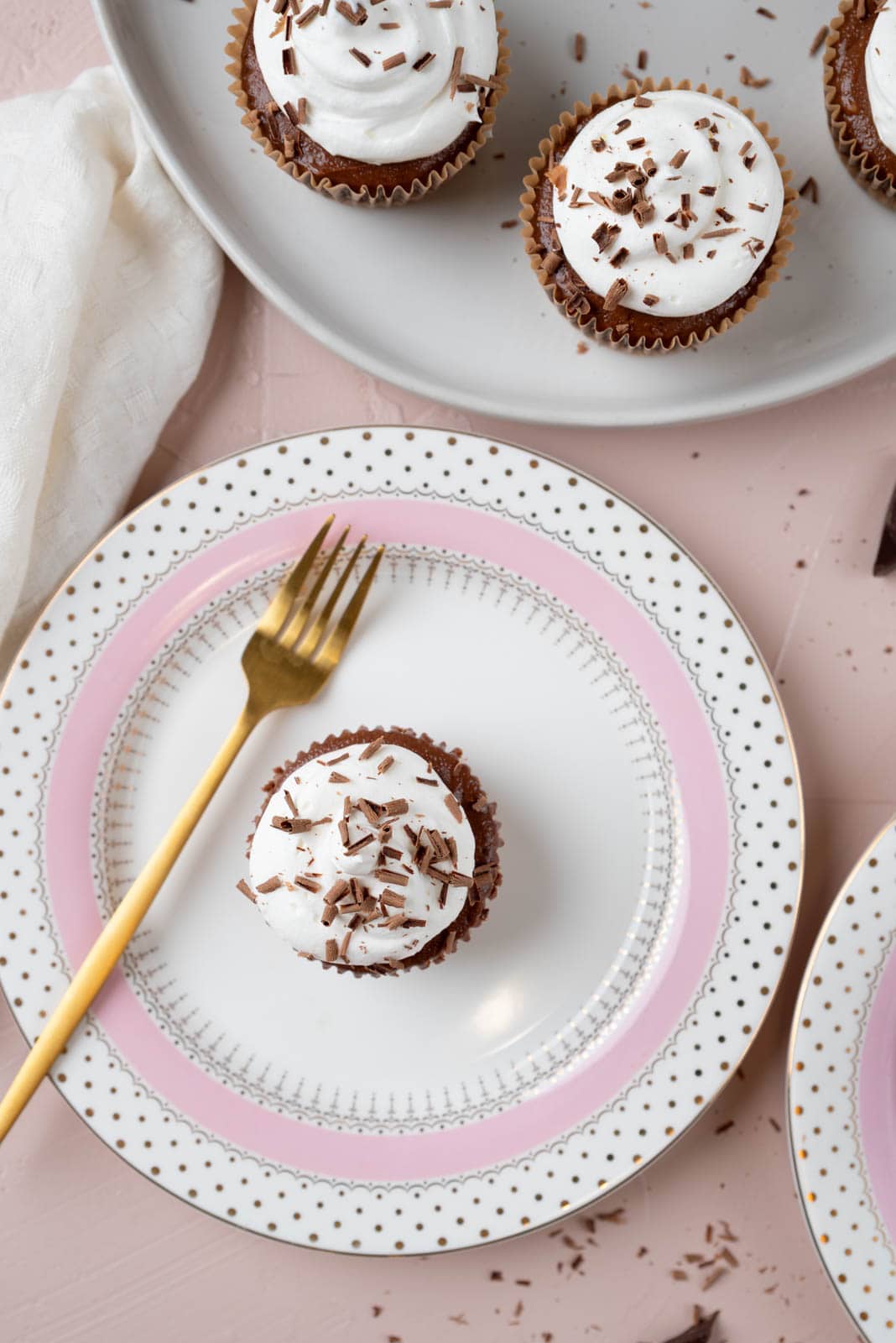 Overhead wide shot of batch of mini chocolate cheesecakes, some on a serving platter and one on a pink and gold dessert plate with a gold fork. A linen napkin and chocolate shavings scattered around the edges.