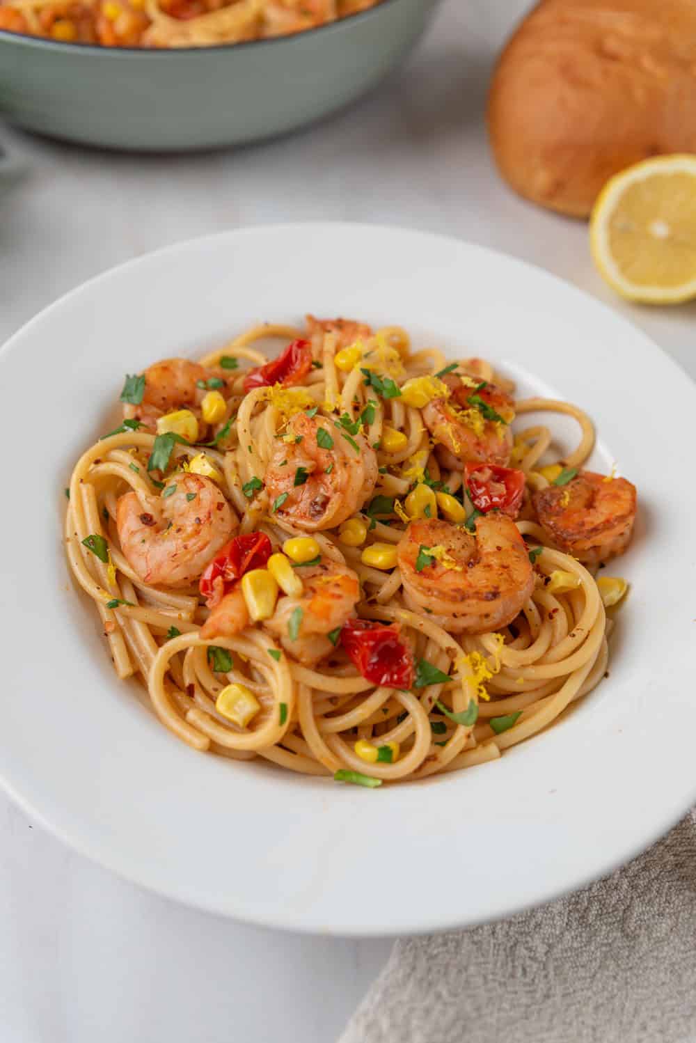A closeup of a white pasta bowl filled with Old Bay Shrimp Pasta. Lemon zest is sprinkled over the top of the dish that is dotted with tomatoes, corn and fresh parsley. A loaf of crusty french bread and the large pasta pot in the background.