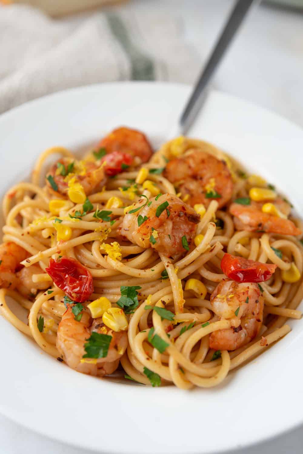 A closeup of a white pasta bowl filled with Old Bay Shrimp Pasta. Lemon zest is sprinkled over the top of the dish that is dotted with tomatoes, corn and fresh parsley. A loaf of crusty french bread and the large pasta pot in the background. A linen napkin rests in the background.