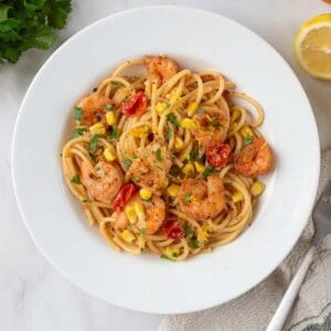 A white bowl filled with pasta. The swirls of pasta are dotted with sear shrimp, cherry tomatoes, corn and fresh parsley. Finally, it is garnished with lemon zest.