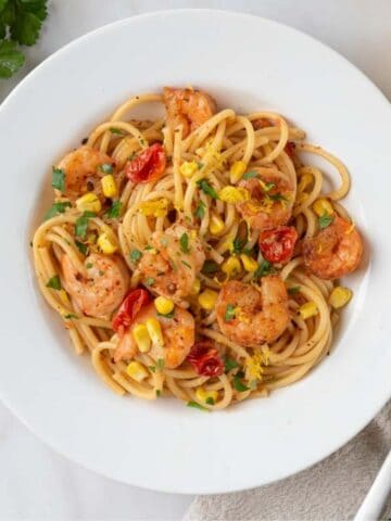 A pasta bowl filled with Old Bay Shrimp Pasta, dotted with corn, cherry tomatoes and fresh parsley. A cut lemon, a green linen napkin, a bundle of parsley and a loaf of crusty bread are on the countertop.