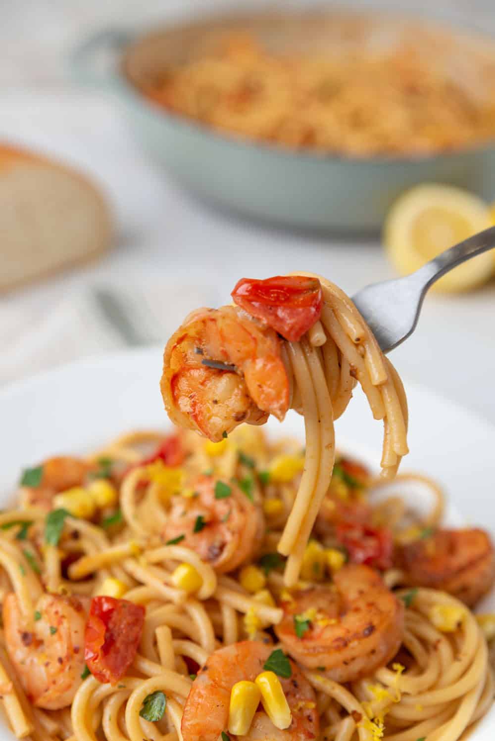 A closeup of a fork holding a swirl of Old Bay Shrimp Pasta with the bowl of pasta resting underneath. The pasta pot, a cut lemon and a loaf of bread out of focus in the abckground.