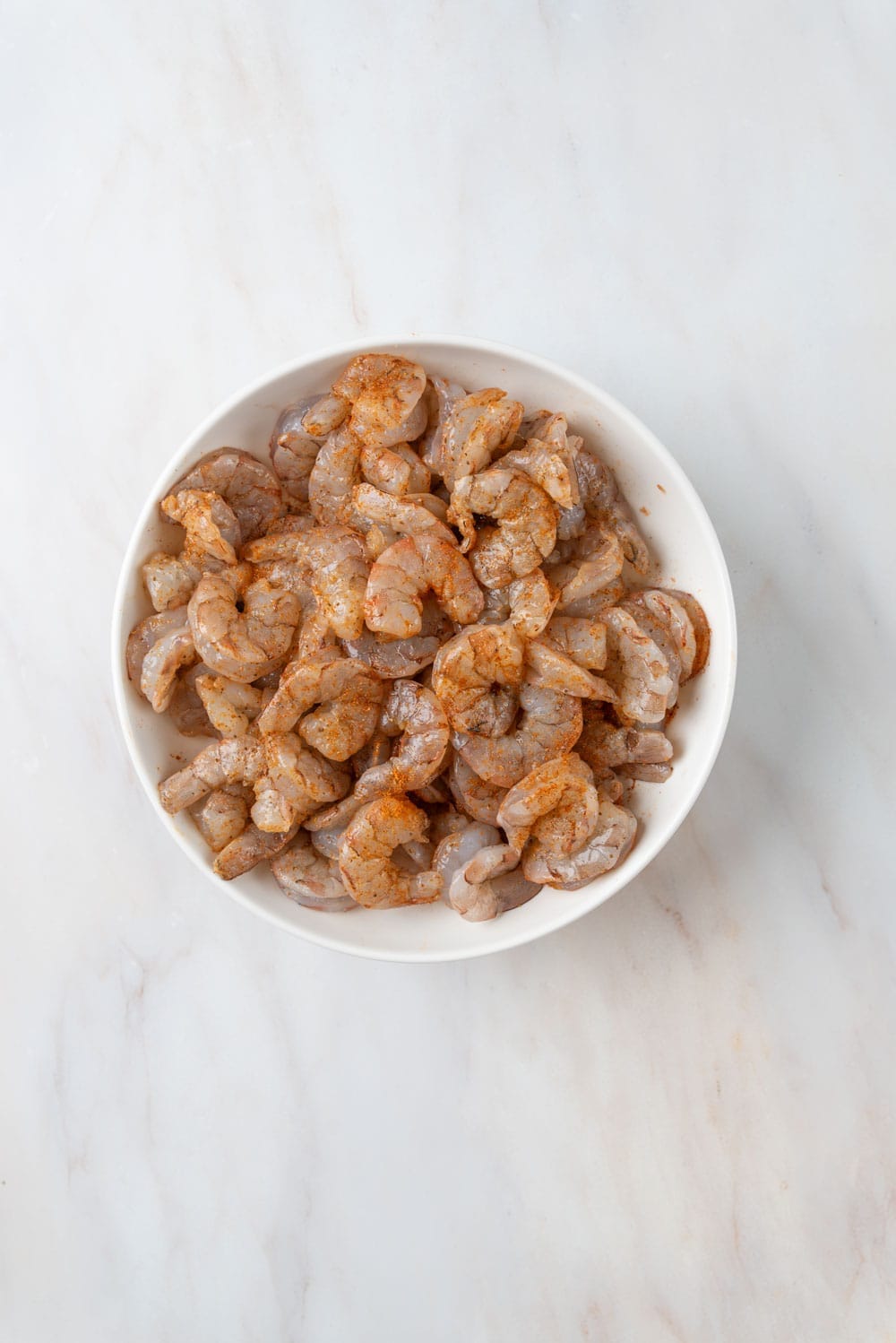 A marble countertop with a white bowl holding raw shrimp, heavily seasoned with Old Bay.