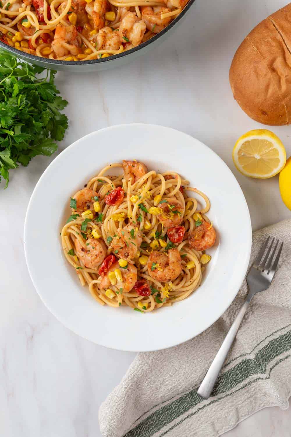 An overhead shot of a white bowl filled with Old Bay Shrimp Pasta. The pasta dish is dotted with seared shrimp, corn, blistered cherry tomatoes and fresh parsley. Near the bowl, on the coutertop rests a linen napkin and fork, a cut lemon, a bundle of parsley, a loaf of bread and the large pasta pot.