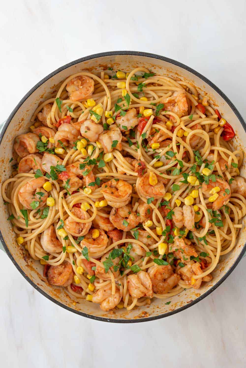 A deep braising pan is filled with swirling piles of Old Bay Shrimp Pasta. Deeply seared pasta, corn and cherry tomatoes dot the pan of pasta with flecks of green from fresh parsley.
