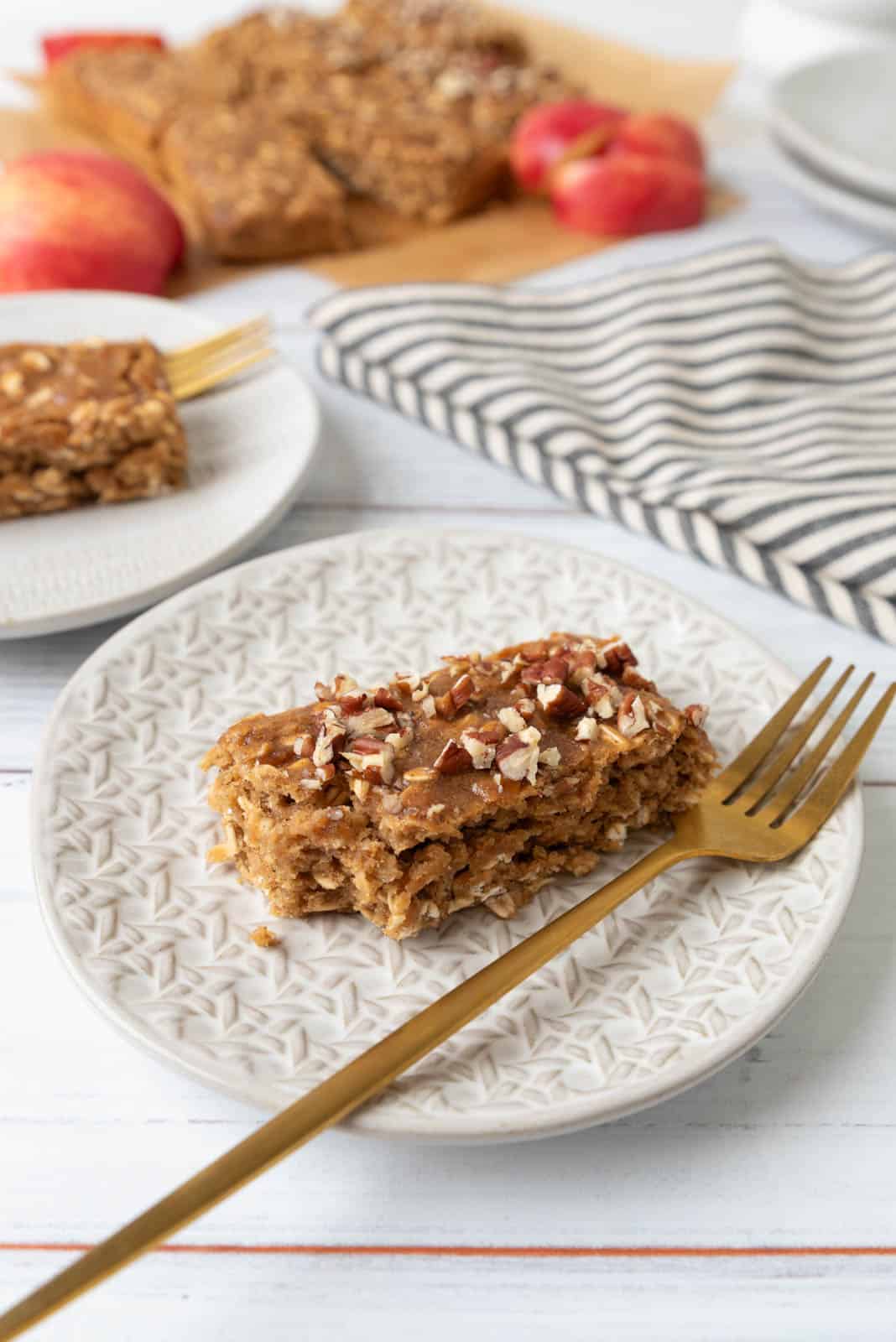 An applesauce oatmeal breakfast bar sprinkled with chopped pecans sitting on a plate with a bite taken from its corner. A gold fork rests on the plate. More bars, another plate, a sliced apple and a striped napkin in the background.