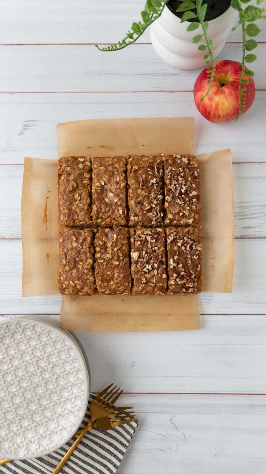 Baked applesauce oatmeal bars cooled and cut on their parchment paper. A stack of plates, napkins and forks sit beside them. A red apple and green plant in the far corner above the bars.