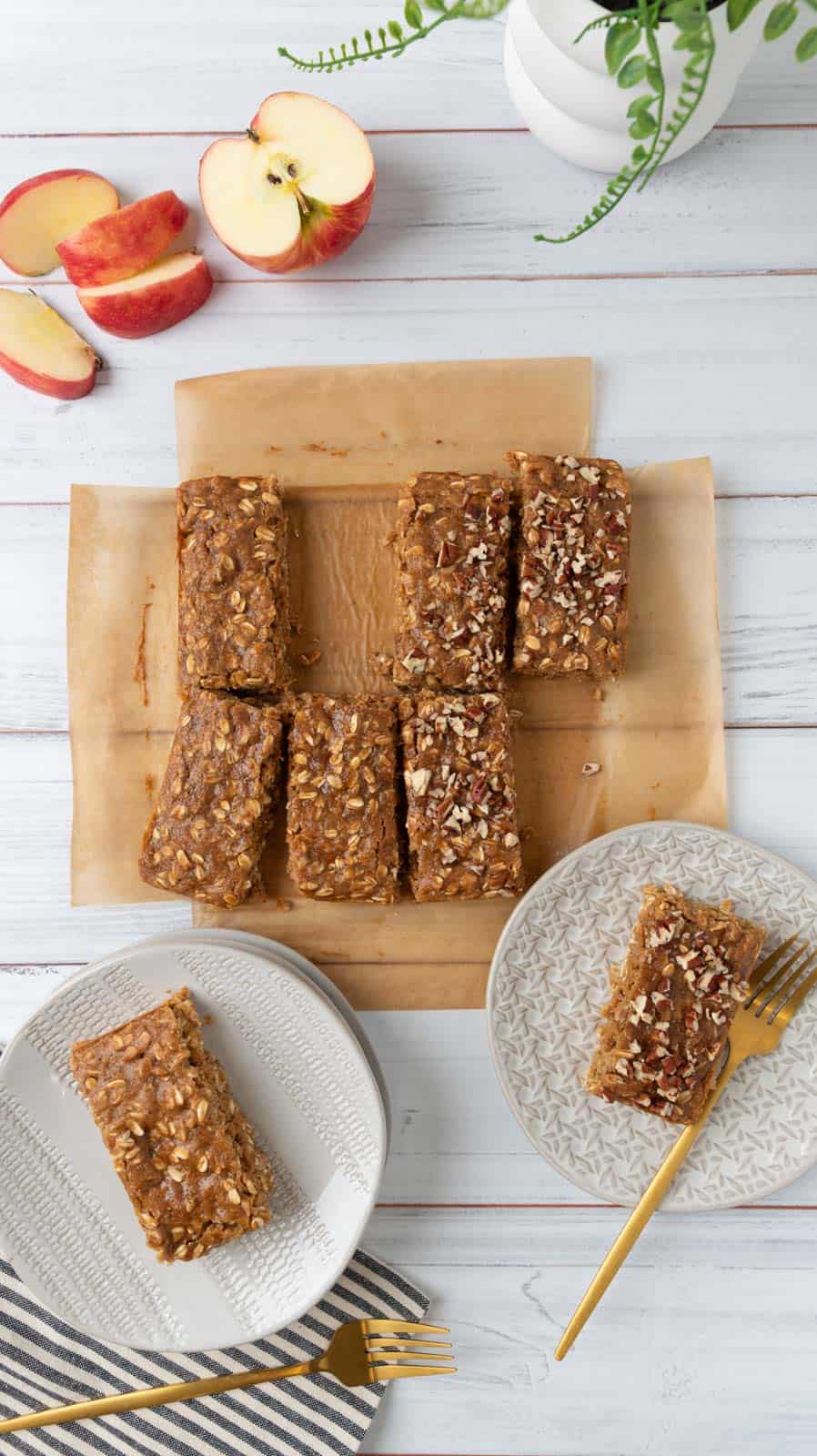 An overhead view of two oatmeal bars sitting on cream plates on a whitewash wood tabletop. The rest of the bars resting on brown parchment near the plates. A sliced apple, a green houseplant, a striped napkin and gold forks are also in the frame.