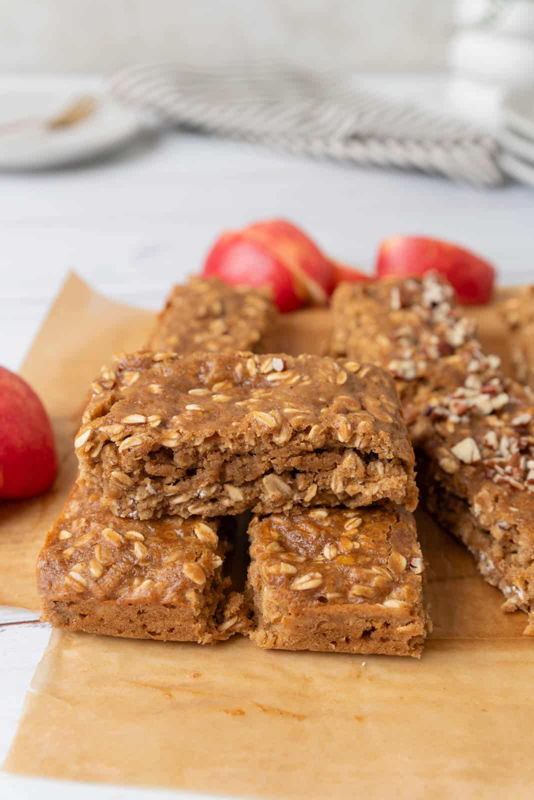 Applesauce oatmeal bars sitting on a piece of brown parchment paper. The bars are stacked in several piles with slices of apple and a striped napkin in the background.