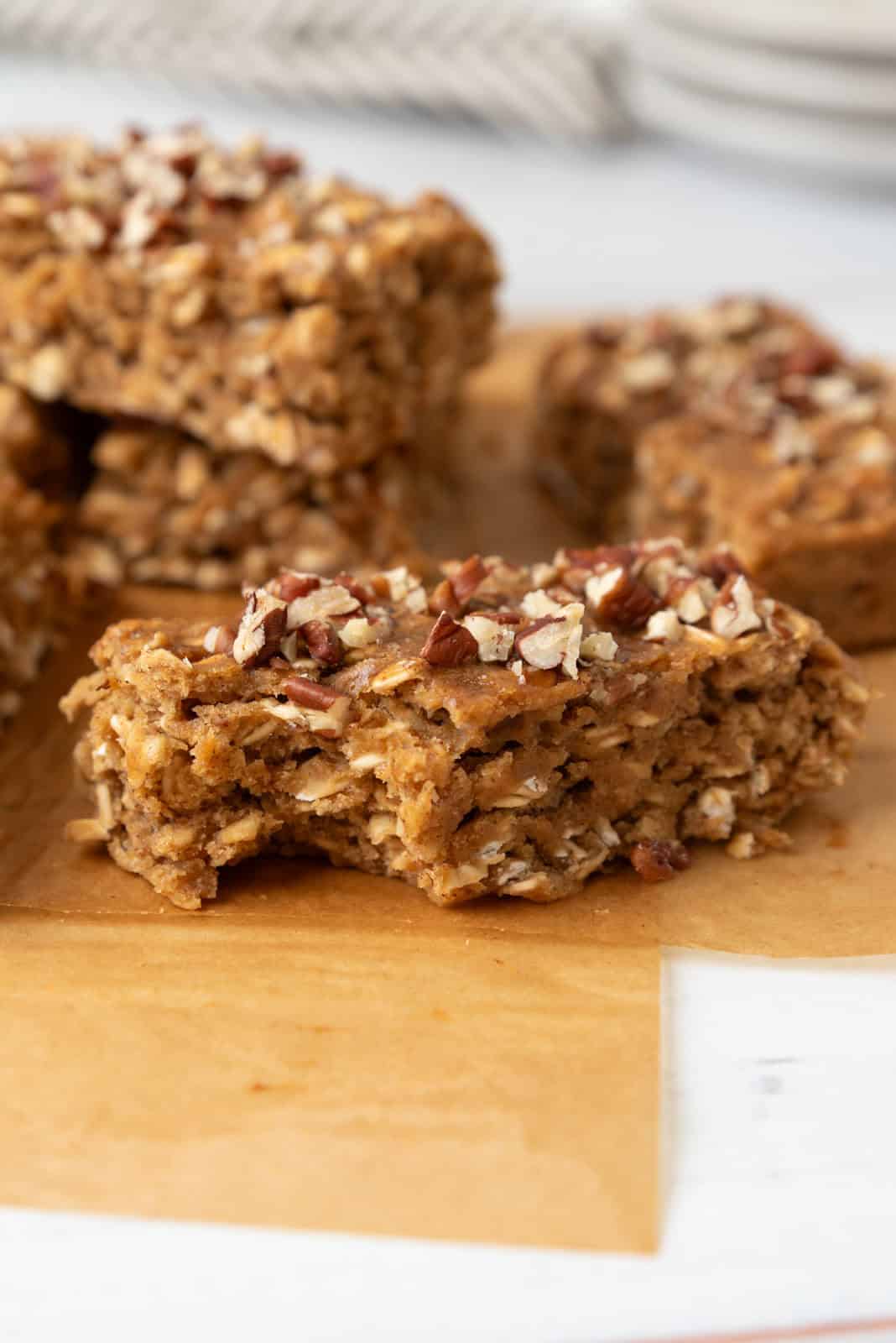 A closeup of a breakfast bar with a bite taken out, resting on parchment paper with more bars stacked in the background.