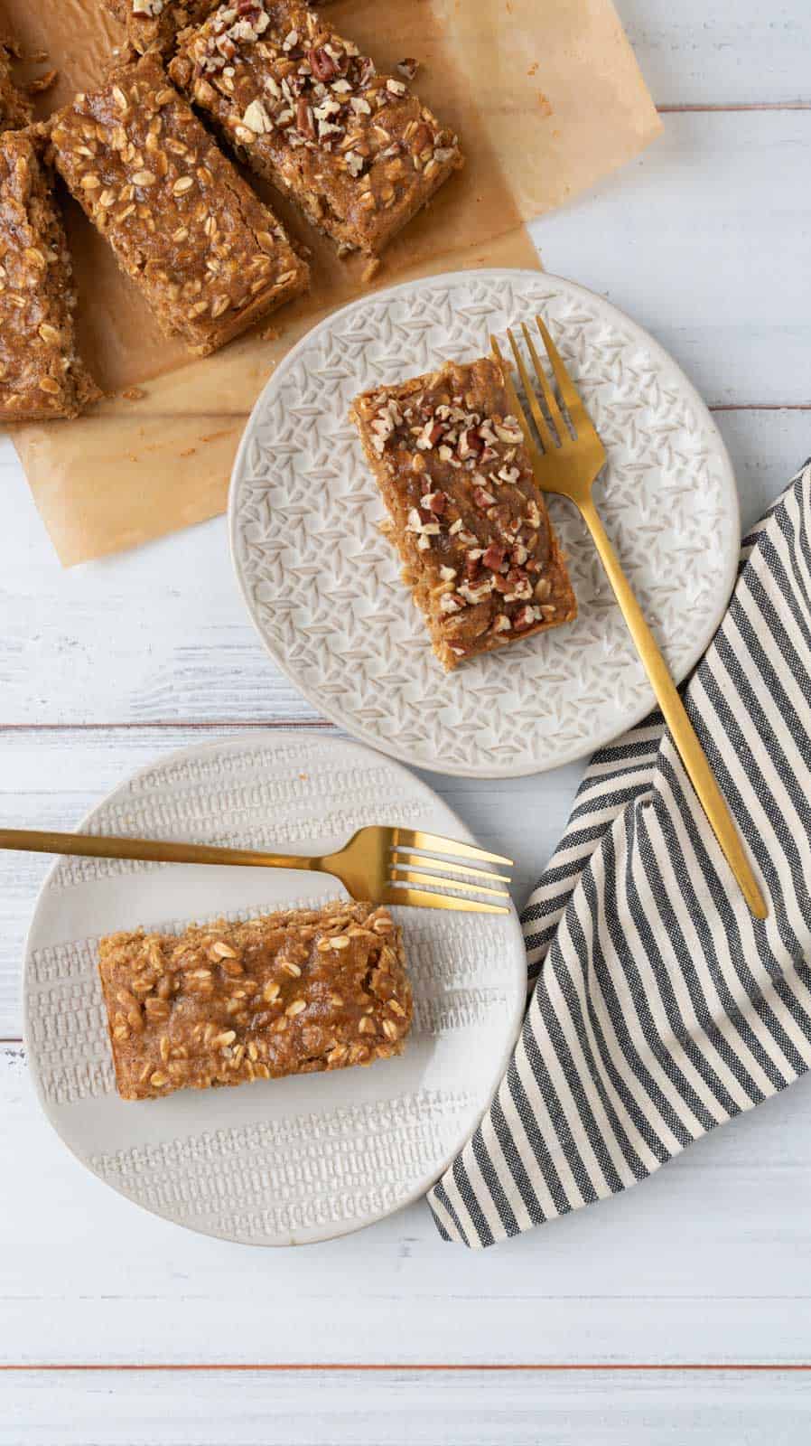 Applesauce oatmeal breakfast bars cut on a sheet of parchment paper with two bars on small cream plates. A black and white striped napkin and gold forks resting on the plates.