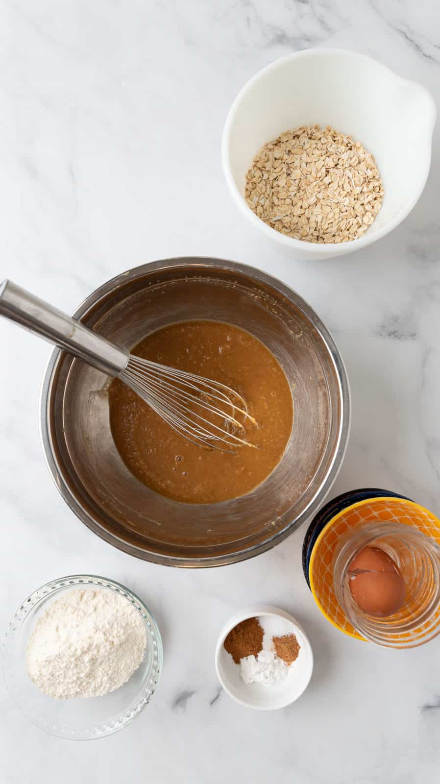 Wet ingredients combined in a stainless steel mixing bowl with a whisk. The dry ingredients in bowls surrounding it.