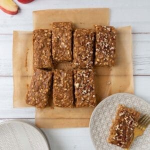 An overhead view of two oatmeal bars sitting on cream plates on a whitewash wood tabletop. The rest of the bars resting on brown parchment near the plates. A sliced apple in the corner.