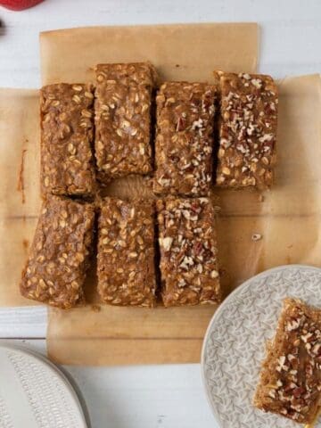 An overhead view of two oatmeal bars sitting on cream plates on a whitewash wood tabletop. The rest of the bars resting on brown parchment near the plates. A sliced apple in the corner.