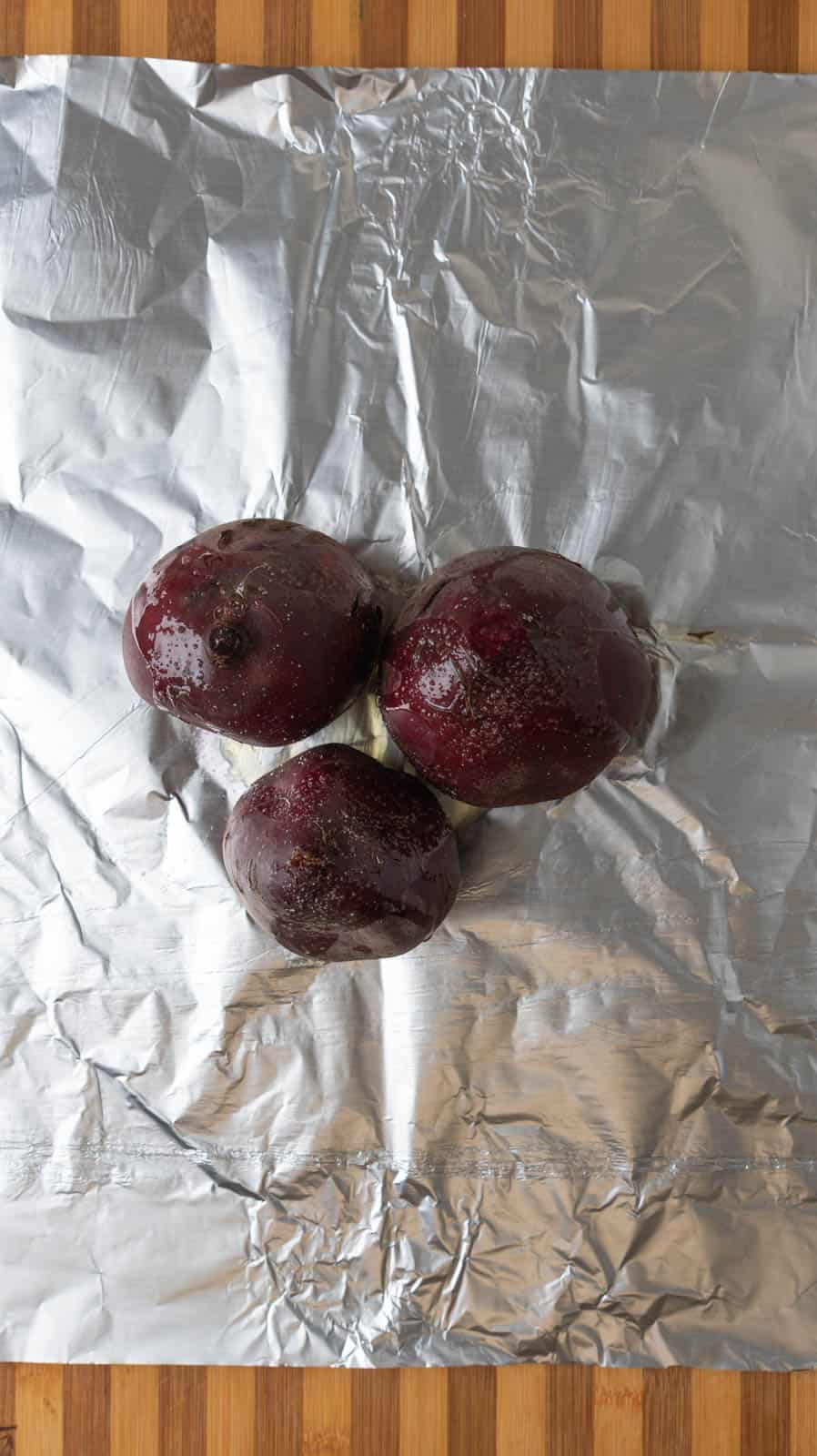 Three large beets drizzled in oil and salt resting on a piece of aluminum foil, ready to be wrapped into a packet and baked.