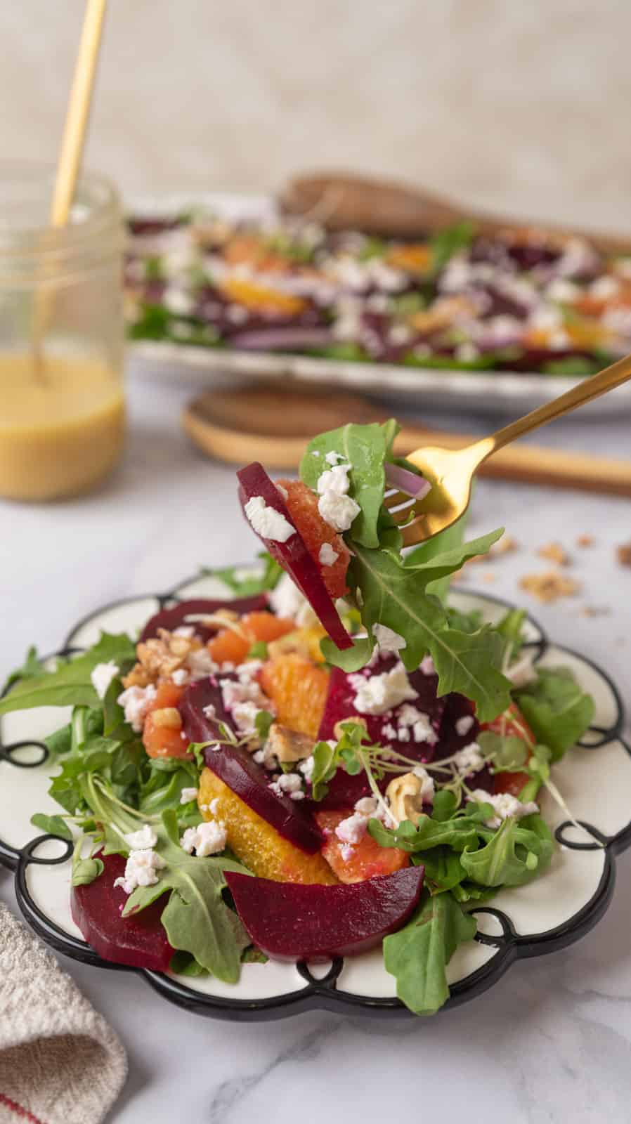A fork full of beet and citrus salad with feta cheese hovering over a plate of salad. A napkin in the corner along with a jar of dressing, wooden salad tongs and a platter of salad in the background.