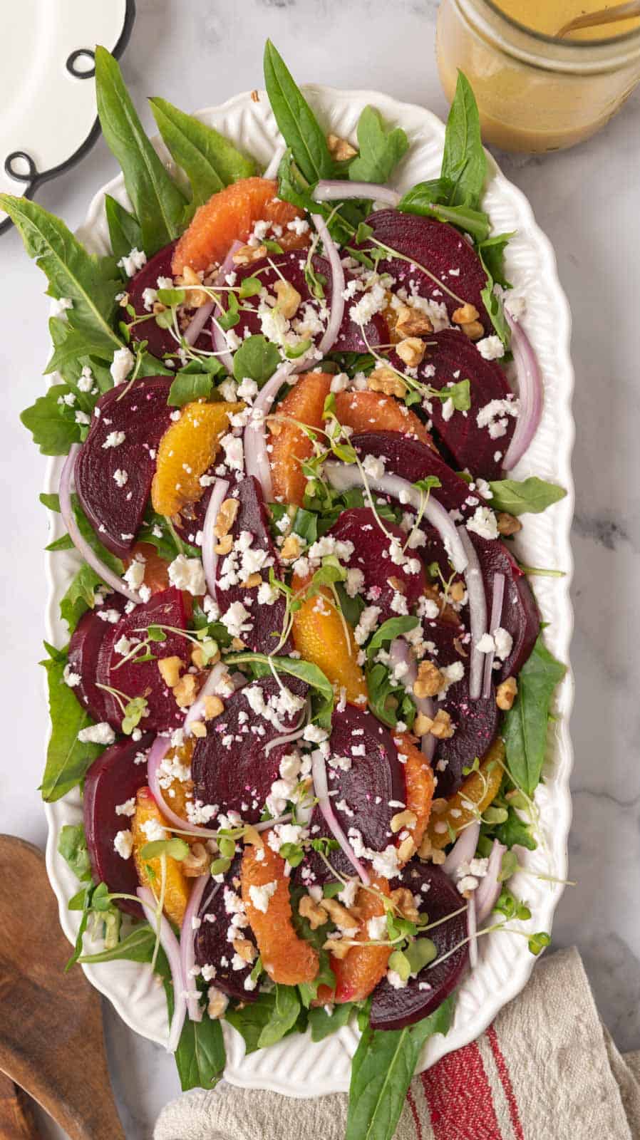 A large white serving platter loaded with a beautiful, colorful beet and citrus salad with feta cheese. Salad plates and a jar of dressing waiting beside the platter.