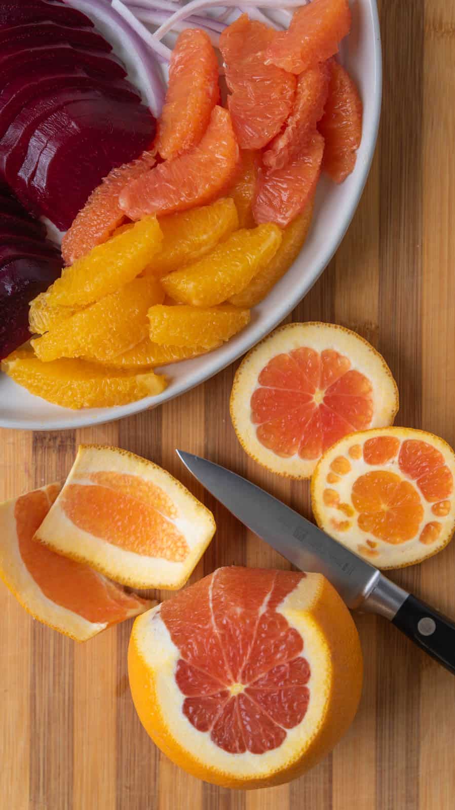 A cutting board holding a plate of segmented oranges while a partially cut orange with a knife sitting near it waits to be finished.