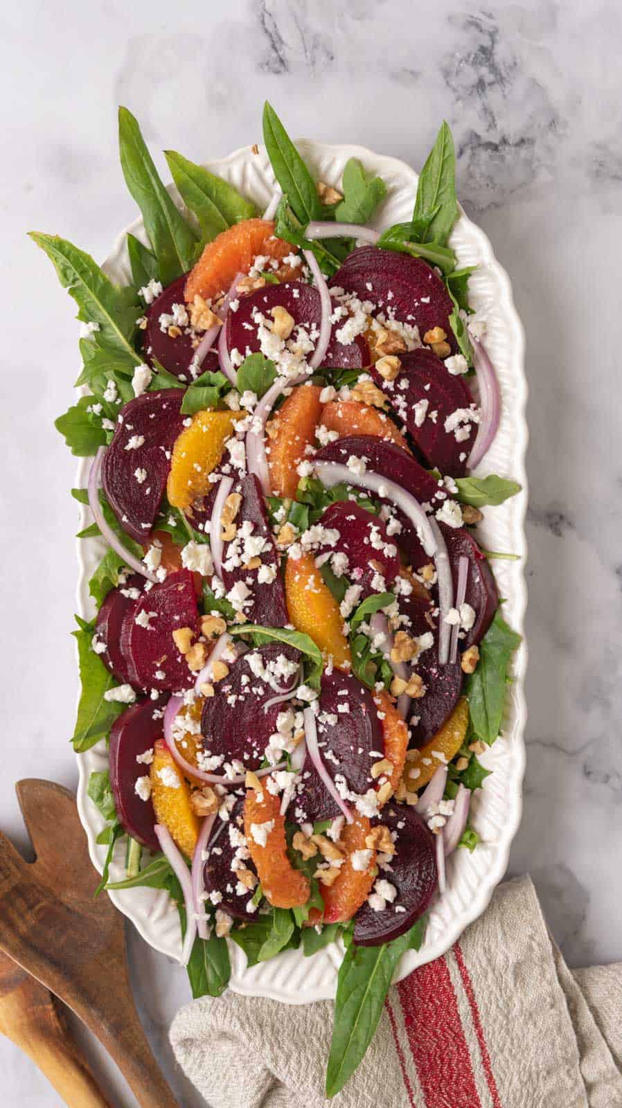 A large white serving platter loaded with a beautiful, colorful beet and citrus salad with feta cheese. Wooden salad tongs and a linen napkin are on the side.