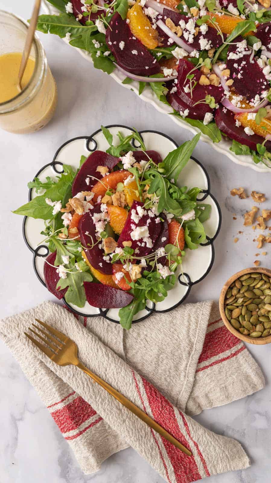 A gorgeous plate of colorful beet and citrus salad with feta cheese sits in the middle with a gold fork and linen napkin resting to the side. Also on the countertop is a small wooden bowl of pepitas, walnuts bits scattered around, a serving platter filled with salad and a mason jar of dressing.