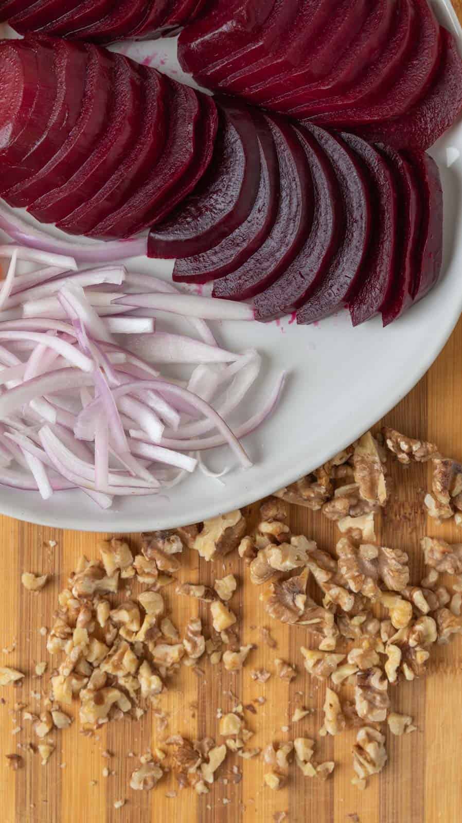 The corner of a plate filled with sliced shallots, sliced roasted beets rests on a cutting board with chopped walnuts on the cutting board.