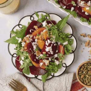 A gorgeous plate of colorful beet and citrus salad with feta cheese sits in the middle with a gold fork and linen napkin resting to the side. Also on the countertop is a small wooden bowl of pepitas, walnuts bits scattered around, a serving platter filled with salad and a mason jar of dressing.