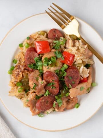 A soft cream plate with a tan rim holding a delicious and colorful plate of chicken dirty rice. A napkin to one side, a fork on the rim of the plate.