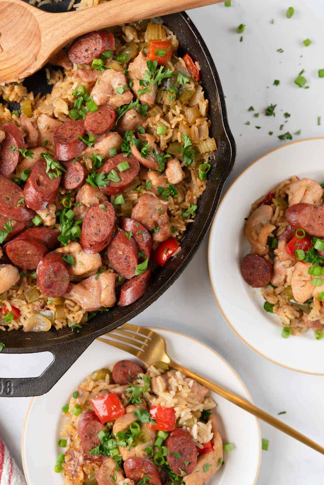 Chicken & Chicken Sausage Dirty Rice plated and ready to serve with the skillet filled with more to the side of the plate. Gold forks resting on the plates and chopped parsley scattered around.
