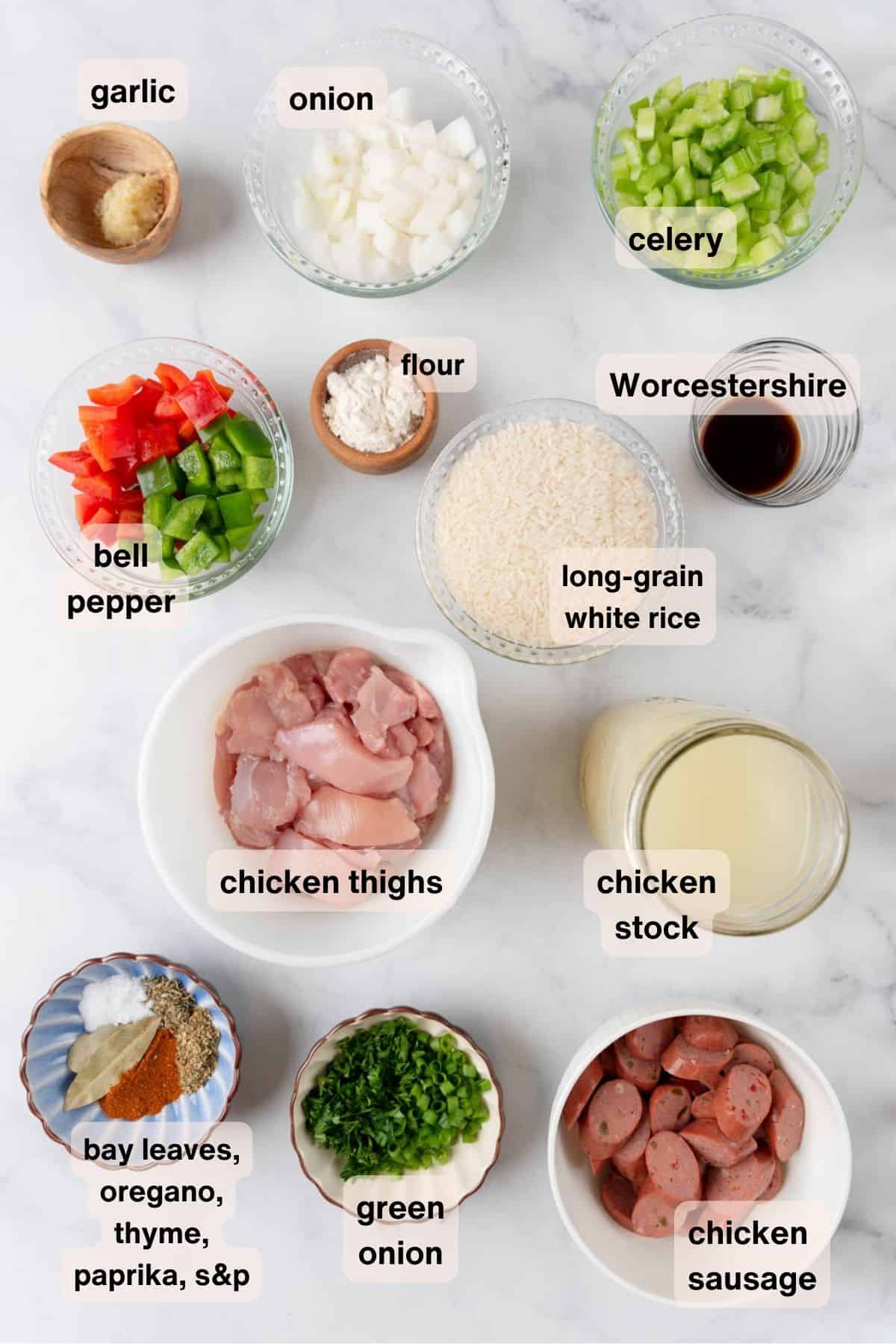 Ingredients for chicken & chicken sausage dirty rice scattered around a white marble countertop in various bowls and dishes.