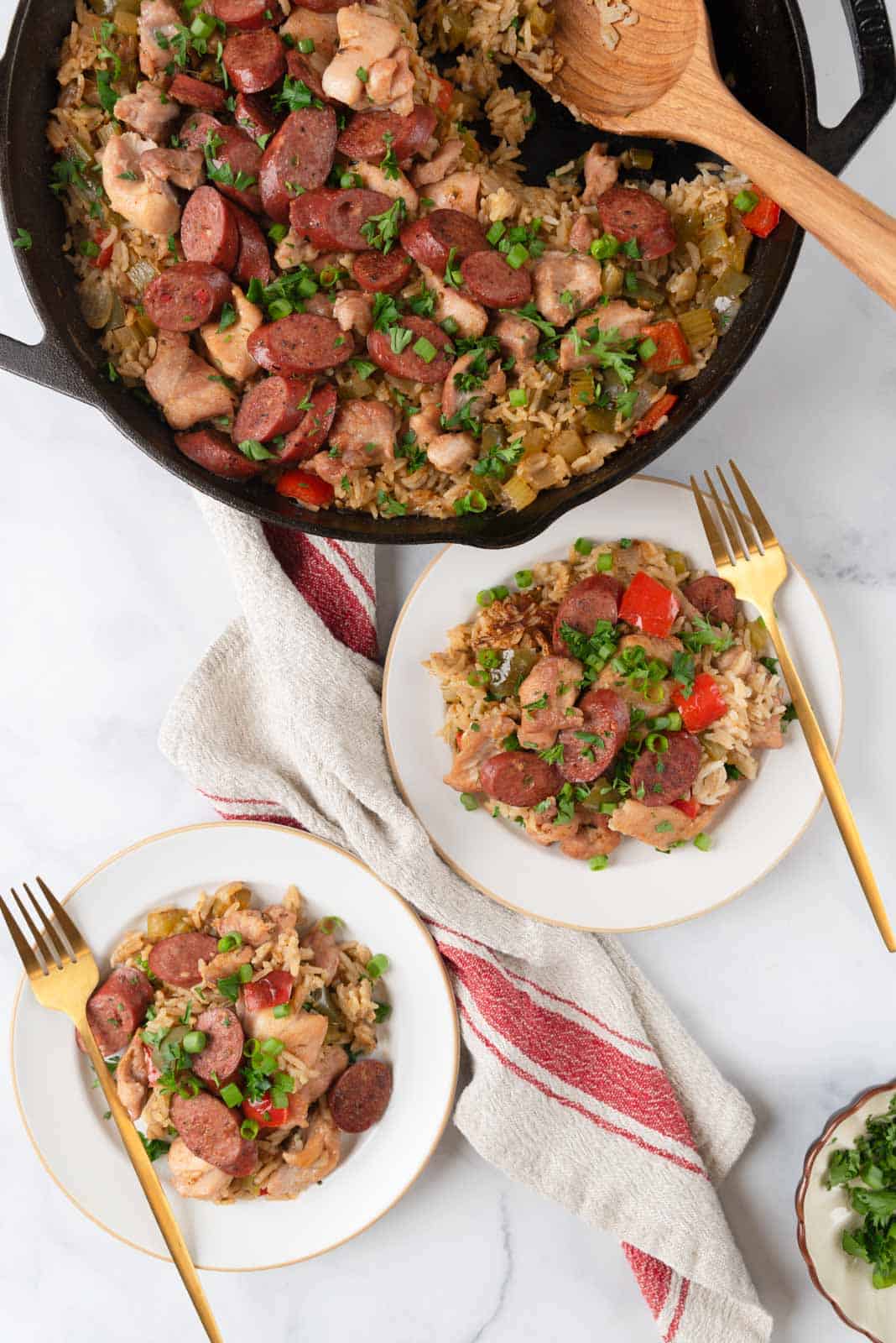 Two plates heaping with chicken dirty rice, gold forks resting on the rims. A big linen napkin on the tabletop with the corner of the cast iron skillet still filled with the dish. A small dish of green onion and parsley ti the side.
