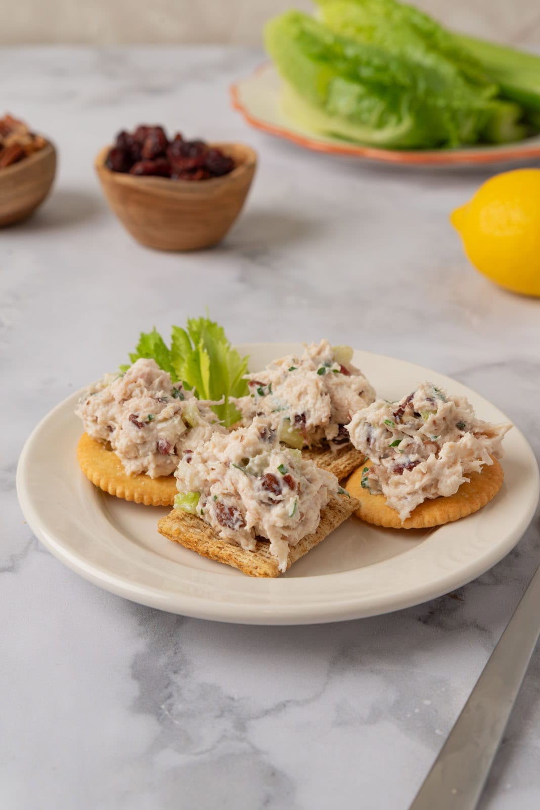 A cream colored plate with crackers topped with cranberry pecan chicken salad. A fork on the white marble countertop to the side of the plate and another plate near with lettuce and celery sticks. A lemon and two small wooden bowls holding dried cranberries and pecans in the background.