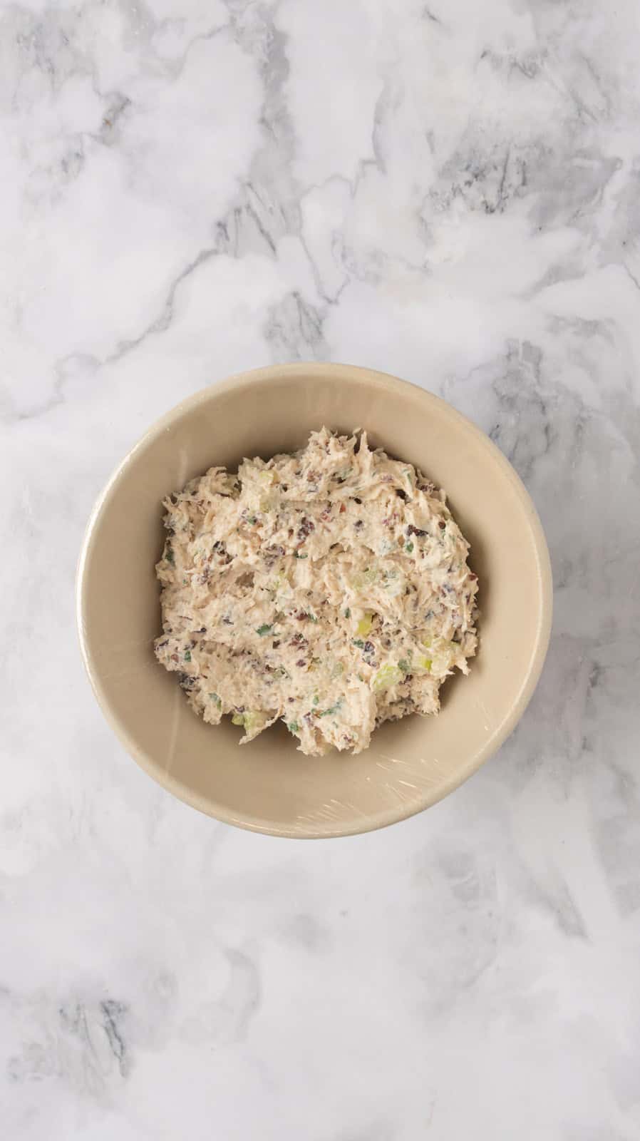 Chicken salad in a mixing bowl, covered in plastic wrap, ready to chill in the refrigerator.