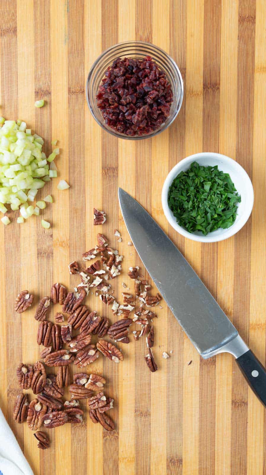 A wooden cutting board with two small bowls holding chopped dried cranberries and shopped parsley. A chefs knife with chopped pecans and a pile of diced celery also on the board.