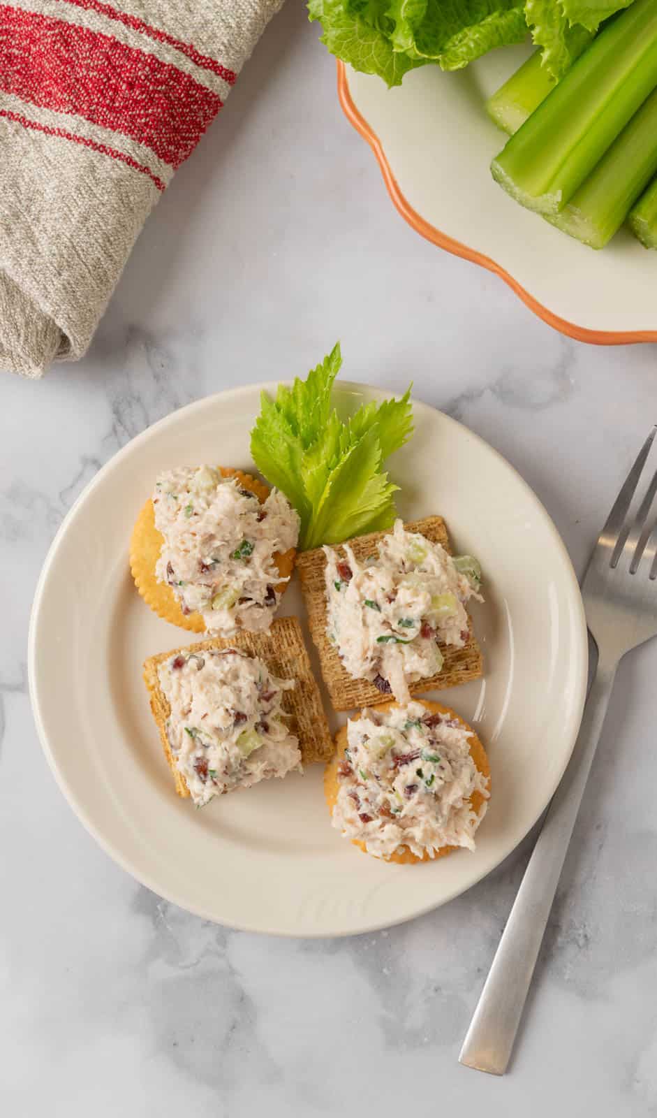 A cream colored plate with crackers topped with cranberry pecan chicken salad. A fork on the white marble countertop to the side of the plate and another plate near with lettuce and celery sticks.