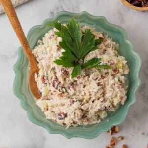 A fluted green glass serving bowl filled with chicken salad, topped with parsley leaves. The white marble countertop also holds a cream plate with crackers topped with cranberry pecan chicken salad, a small wooden bowl with dried cranberries. The countertop is scatter with chopped pecans.