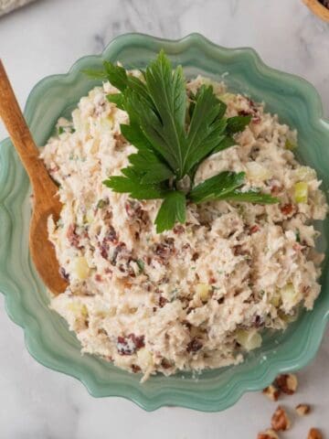 A fluted green glass serving bowl filled with chicken salad, topped with parsley leaves. The white marble countertop also holds a cream plate with crackers topped with cranberry pecan chicken salad, a small wooden bowl with dried cranberries. The countertop is scatter with chopped pecans.