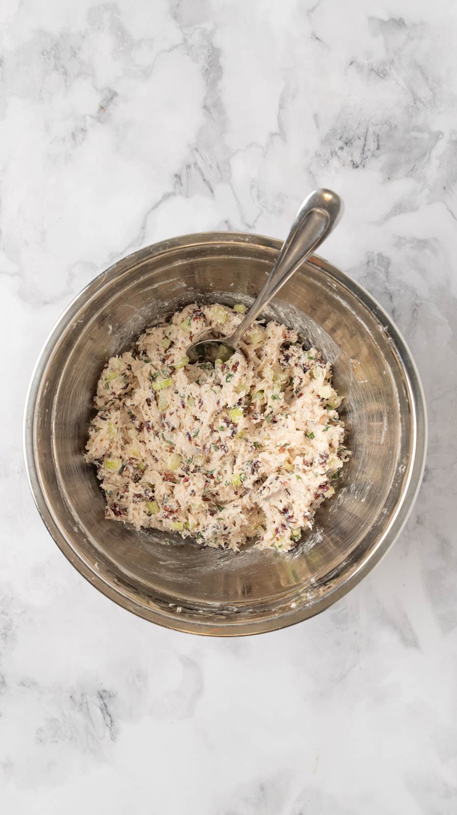 Chicken salad mixed together in a stainless steel mixing bowl on a white marble countertop.
