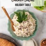 A fluted green glass serving bowl filled with chicken salad, topped with parsley leaves. The white marble countertop also holds a cream plate with crackers topped with cranberry pecan chicken salad, a small wooden bowl with pecans and platters with croissants and lettuce leaves.