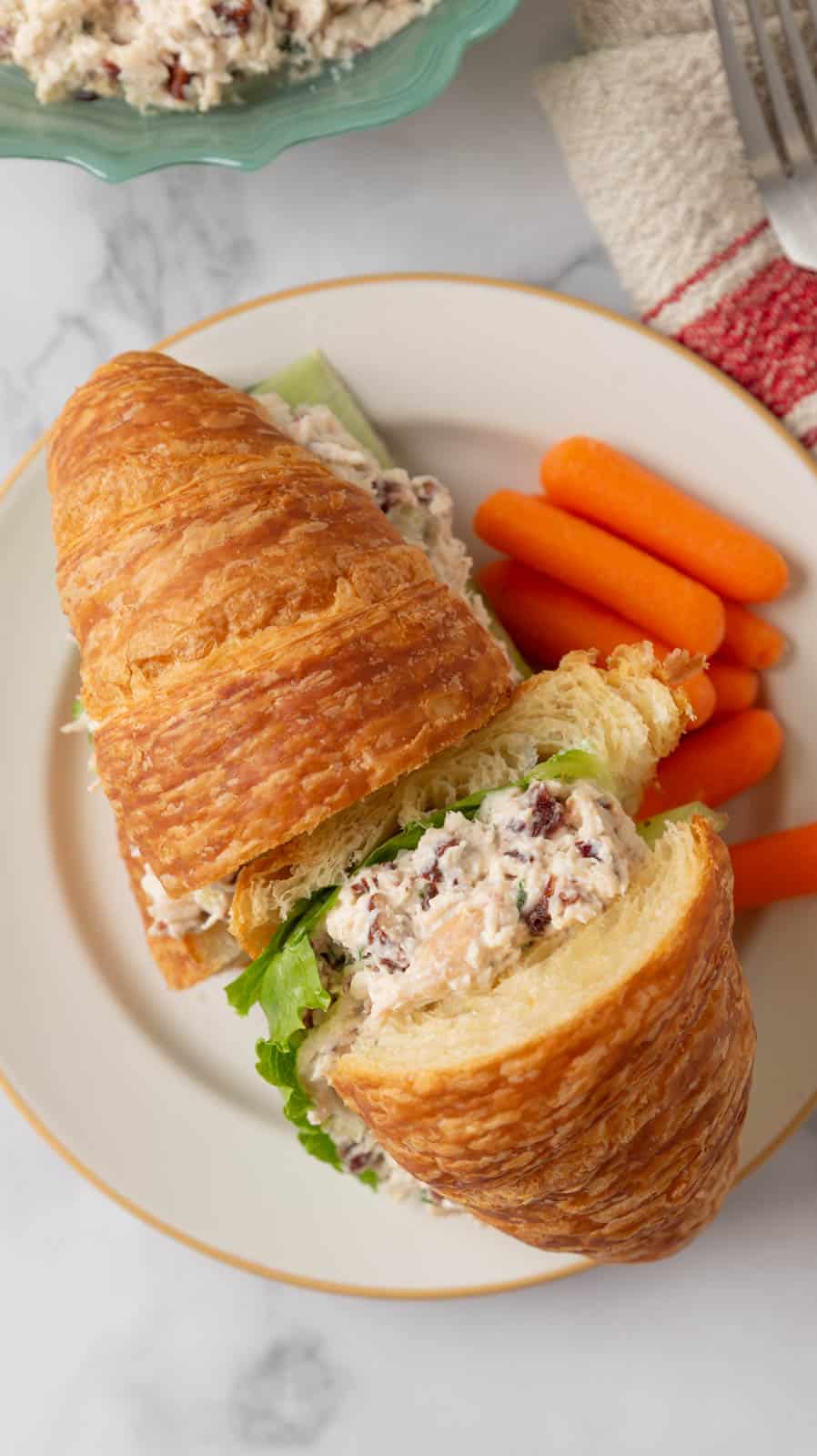 A cranberry pecan chicken salad croissant sandwich with baby carrots on a cream plate.