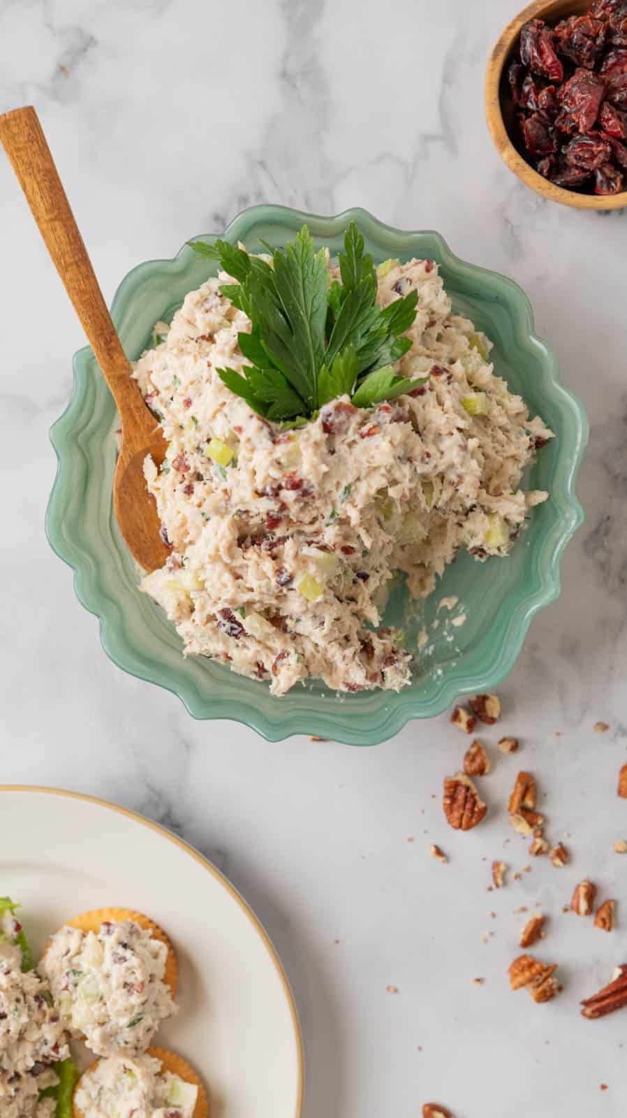 A fluted green glass serving bowl filled with chicken salad, topped with parsley leaves. The white marble countertop also holds a cream plate with crackers topped with cranberry pecan chicken salad, a small wooden bowl with dried cranberries. The countertop is scatter with chopped pecans.