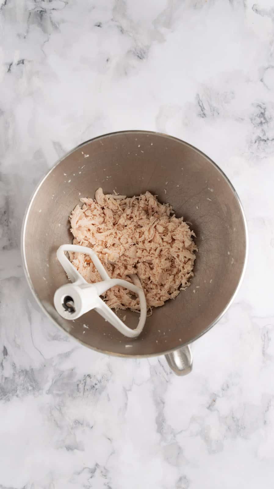 Cooked chicken breasts shredded in a mixing bowl with a paddle attachment resting in the bowl.