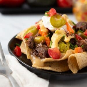 A close-up of a small black plate loaded with cheesy nachos sitting on a white marble countertop with a napkin and fork beside the plate.